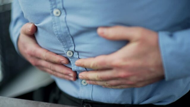 Close up. Male hands holding massaging stomach. Businessman in a blue shirt feels stomach pain sitting at workplace in business office. Sick man suffers from bloating, gastritis poisoning or spasms
