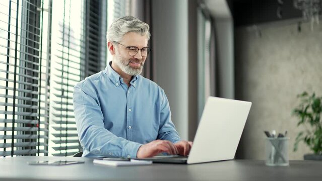 Mature gray haired bearded businessman works on a laptop while sitting at workplace in business office. Senior entrepreneur typing massage chatting banking in computer app, using software application