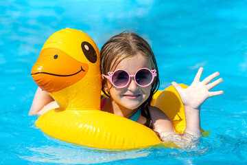 A little girl in sunglasses swims in an outdoor pool hugging an inflatable duckling. Sea holidays on vacation. Pretty female child smiles and swims along with inflatable yellow duck-shaped circle. - obrazy, fototapety, plakaty