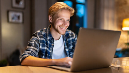 Smiling young man using laptop at home sitting at table at night