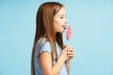 Portrait of positive little girl wearing casual clothes, holding lollipop candy, looking at it,...