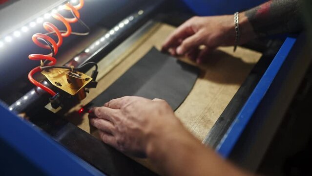 Close up shot of leather worker putting piece of black leather on laser engraving machine. Craftsman working with material square cut to make product