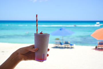 Close up of a cold cocktail drink  with a straw and a cherry on the top held in hand. Blurry beach...