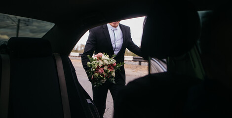 A handsome, young, smiling groom in a black suit stands near a black decorated car with a bouquet...