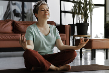 A middle aged lady is practicing yoga at home. A woman in positive mood with closed eyes and slight...