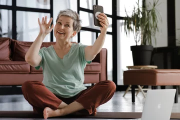 Tuinposter A middle aged lady practices yoga in the large hall of the house. A smiling woman sits on a mat on the floor in the lotus position, has a break time from training, uses a smartphone for video © CinemaF
