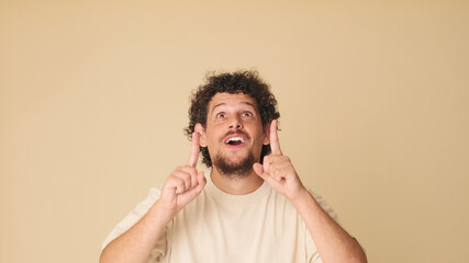 Guy with curly hair dressed in beige t-shirt pointing with fingers to show place for mockup, showing advertisement, advertising sign or announcement with copy space on beige background in studio 