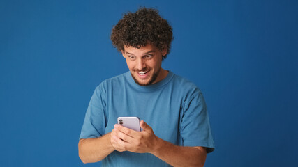 Amazed guy with curly hair dressed in blue t-shirt, holding mobile phone, shopping online, wow effect on blue background in the studio