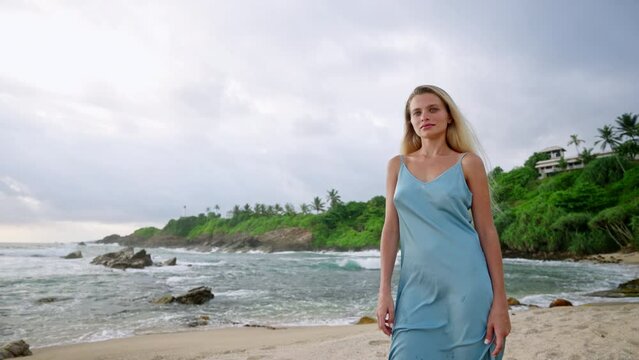 Elegant woman strolls on tropical beach in blue dress, sun-kissed skin gleaming. Luxurious vacation, resort wear showcase, serene nature backdrop. Model exudes high-end fashion, summer vibes.