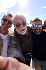 Three middle-aged Caucasian men wearing sunglasses taking a selfie with a cell phone, happy and smiling looking at camera enjoying their vacation outdoors on a sunny day. Vertical photography.