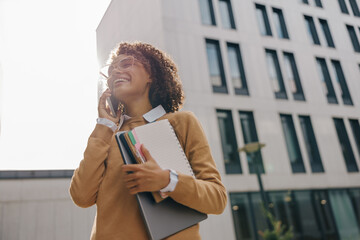 Pretty female entrepreneur is talking phone while standing with laptop on modern building background
