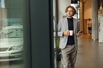 Handsome male office worker use mobile phone and drink coffee while standing near window
