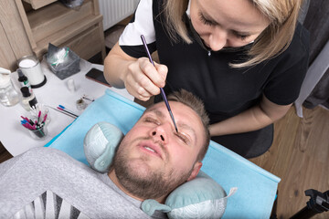Obraz na płótnie Canvas A handsome young man with his eyes closed lies on an eyelash styling procedure.The cosmetologist applies a lamination product to the eyelashes with a brush.Cosmetology for men, professional skin care