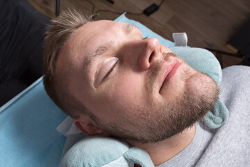 A handsome young man with closed eyes and unshaven stubble lies on the procedure of eyelash...