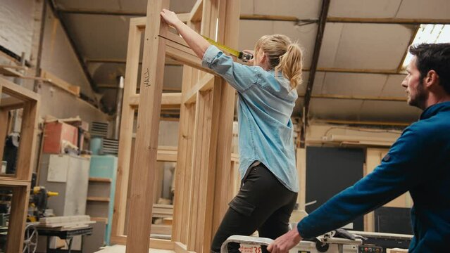Male and female carpenters measuring piece of wood on window frame in workshop using retractable metal ruler - shot in slow motion