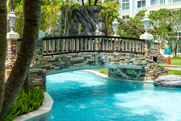 A stone bridge with railings and a fountain spans a blue pool in a condominium at a tropical resort in sunny weather.