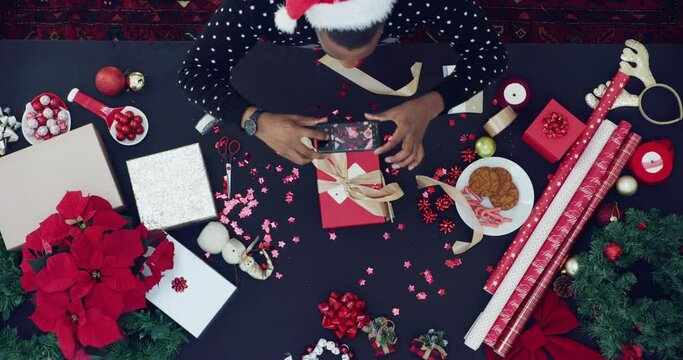 Christmas, gift and person with phone, wrapping paper and santa hat with decoration on table from above. Festive photography, smartphone and man with present, memory and celebration of holiday giving