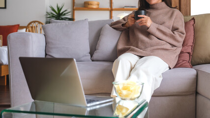 Closeup image of a young woman drinking coffee and eating potato chips while watching on laptop...