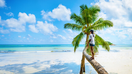Young woman at a palm tree on a white tropical beach with turquoise colored ocean Anse Volbert...