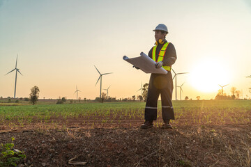 Male engineer in uniform wearing helmet stands and monitors wind turbine station operation holding blueprints using renewable energy until sunset