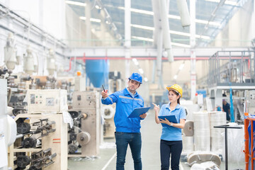 in large factories Asian female supervisor and European male engineer Pointing to a working plastic and steel production machine, wearing a uniform, hard hat, holding a note, a radio.