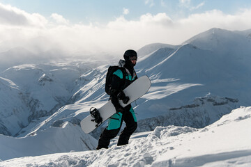 Snowboarder climbs a snowy mountain on foot, holding his snowboard in his hands - obrazy, fototapety, plakaty