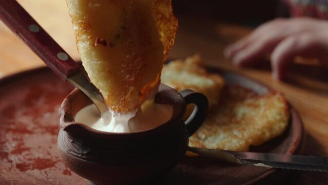 Unrecognizable child dips greasy hash browns into creamy sauce, close-up. Ethnic slavic clay sauce boat with sour cream in cafe. Harmful high calorie fried foods.