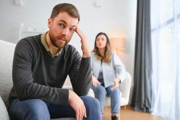 Couple arguing. Wife shouting to her desperate husband sitting on a couch in the living room at home