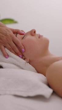 Therapeutic facial massage treatment for young beautiful woman in a medical massage salon. Massage therapist is doing stroking and pushing movements in this scene, side view. Vertical footage.