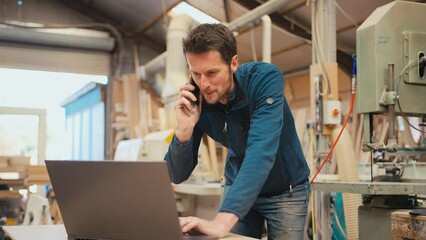 Male carpenter working in woodwork workshop talking on mobile phone whilst using laptop - shot in slow motion - Powered by Adobe