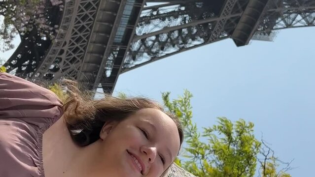 young beautiful girl in a blue dress sits near the Eiffel Tower near a green tree she straightens her braids looks around she is happy and very happy. High quality 4k footage