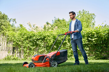 Handsome male gardener in gloves trimming turf with electric mower in garden at day. Side view of smiling bearded guy in denim outfit using lawn trimmer, while working outdoors. Concept of gardening.