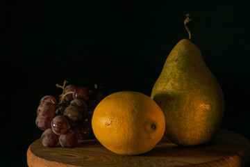 Pear, lemon and grapes on a round wooden table. dark background. A beautiful and unusual combination of fruits in a still life.