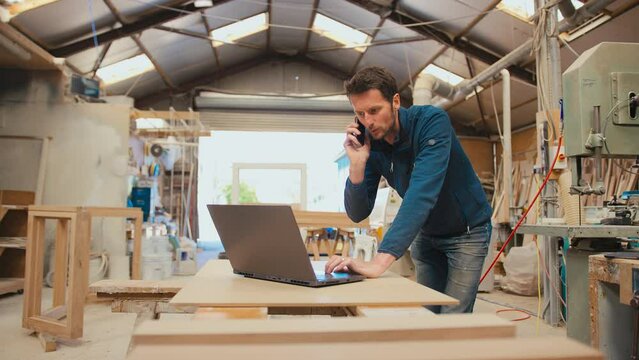 Male carpenter working in woodwork workshop talking on mobile phone whilst using laptop - shot in slow motion