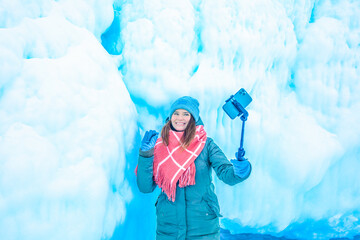 Cheerful female wonderer waves hand and talking via smartphone during video call while standing against big ice rock during winter vacation abroad 