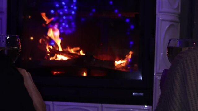 Young couple nearby fireplace sitting and clink wine glasses with champagne. New year or xmas celebration. Indoor shoot with unrecognizable people.
