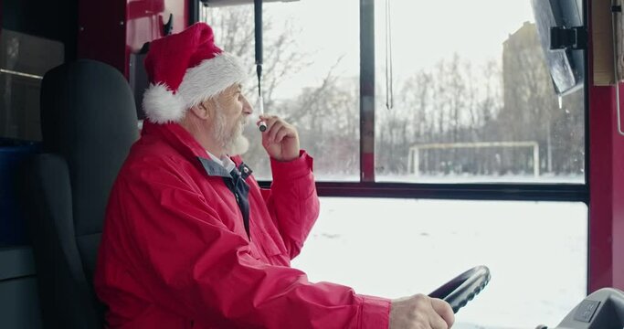Side view of bus driver in Christmas hat sitting, speaking in microphone. Old man with grey hair holding hands on helm, looking forward. Concept of urban life and vacation.