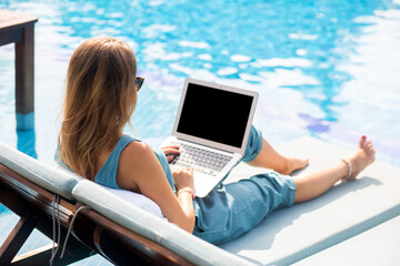 A person is relaxing by a bright blue pool on a sunny day while working or browsing on a laptop....