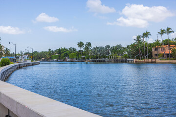 Scenic waterfront promenade in Miami Beach with luxury villas, yacht parking, palm trees, and a backdrop of blue sky and white clouds.