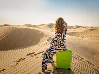 Arabian woman in stylish zebra suit with bright light green or yellow suitcase in sands. Concept and idea of travel to United Arab Emirates, sand dunes in desert of Dubai at sunset. - obrazy, fototapety, plakaty