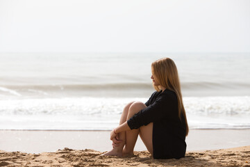 Beautiful young blonde woman in black shirt sits on the shore of the beach sad and depressed. The...