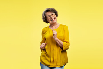 Smiling senior 70s woman wearing casual clothes and eyeglasses dancing isolated on yellow background