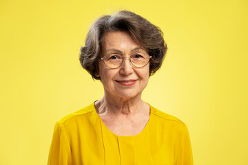 Attractive smiling senior woman, happy grandmother wearing eyeglasses and casual yellow blouse, looking at camera standing isolated on yellow background. Retired concept