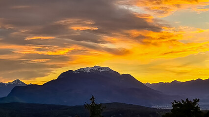 Scenic view of snow capped mountain peak Dobratsch at sunset seen from Taborhoehe in Carinthia, Austria, Europe. Sky has vibrant orange and pink colors with clouds swirling around summit. Serenity