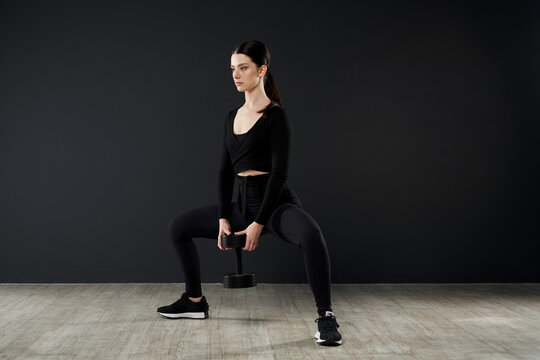 Sporty young woman in black outfit doing squats with dumbbell during crossfit. Side view of beautiful brunette female with ponytail exercising with additional weight, copy space. Concept of workout.