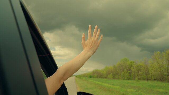 Free girl waves her hand from car window, travels, catches wind with her fingers, Clouds storm. Young female driver plays catches fresh wind with her hand from car window, Road trip on way to vacation