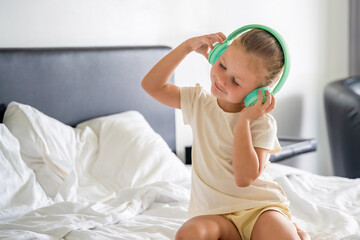 Little girl emotional enjoying music using green kids headphones in home bed