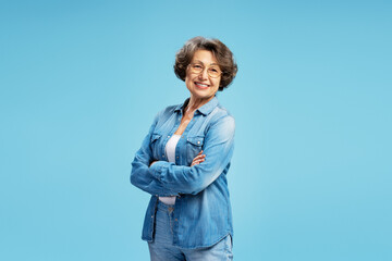 Smiling beautiful senior elderly woman wearing glasses, stylish blue shirt with arms crossed, standing isolated on blue background. Pensioner concept