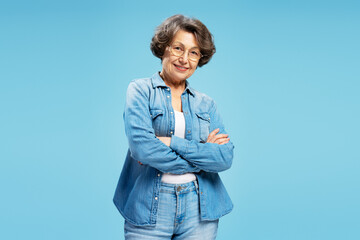 Portrait of confident senior woman wearing eyeglasses and stylish denim shirt, arm crossed isolated on blue background. Advertisement concept - Powered by Adobe