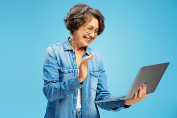 Portrait of smiling gray haired senior woman wearing eyeglasses using laptop, having video call, standing isolated on blue background. Technology concept